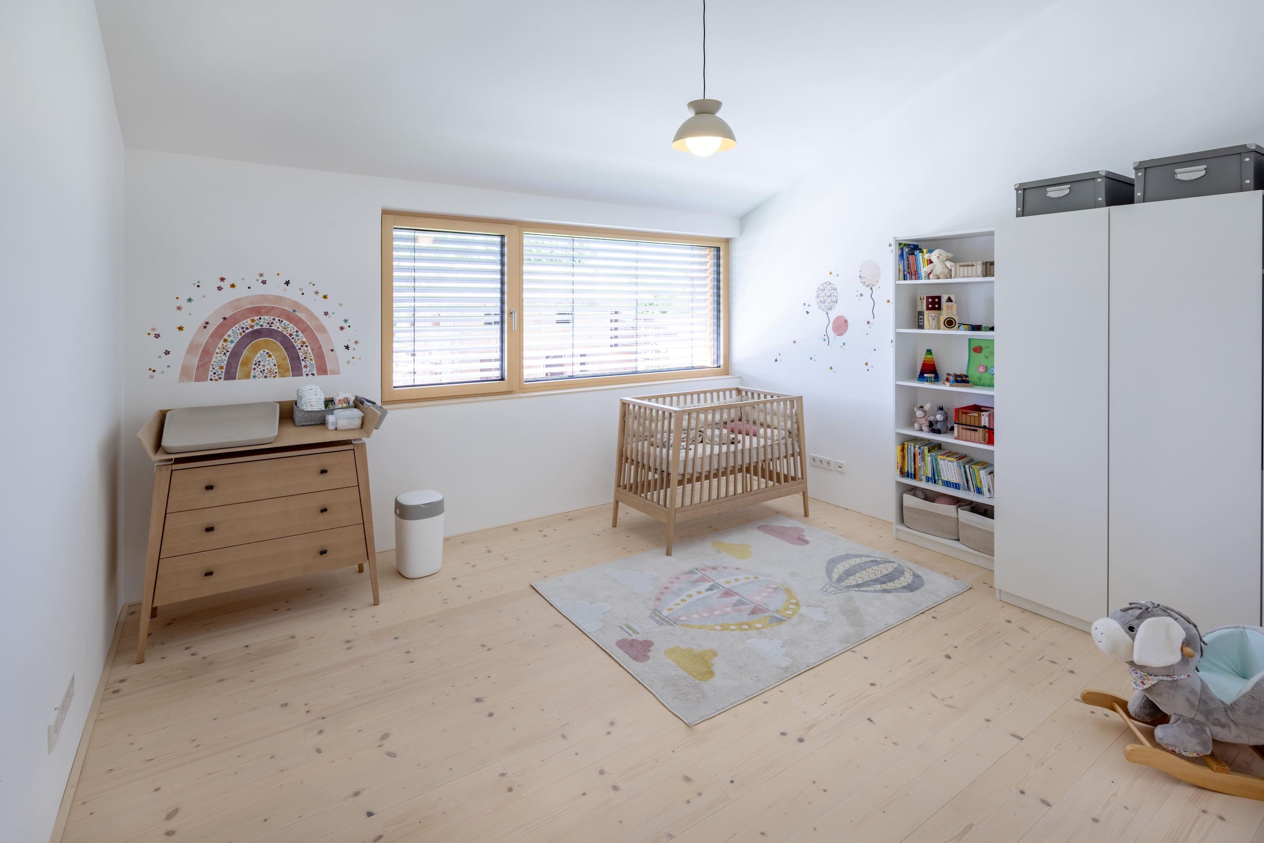 Ein modernes Kinderzimmer mit Holzmöbeln, einem bunten Regenbogen an der Wand und einem Schaukelpferd auf dem Boden.