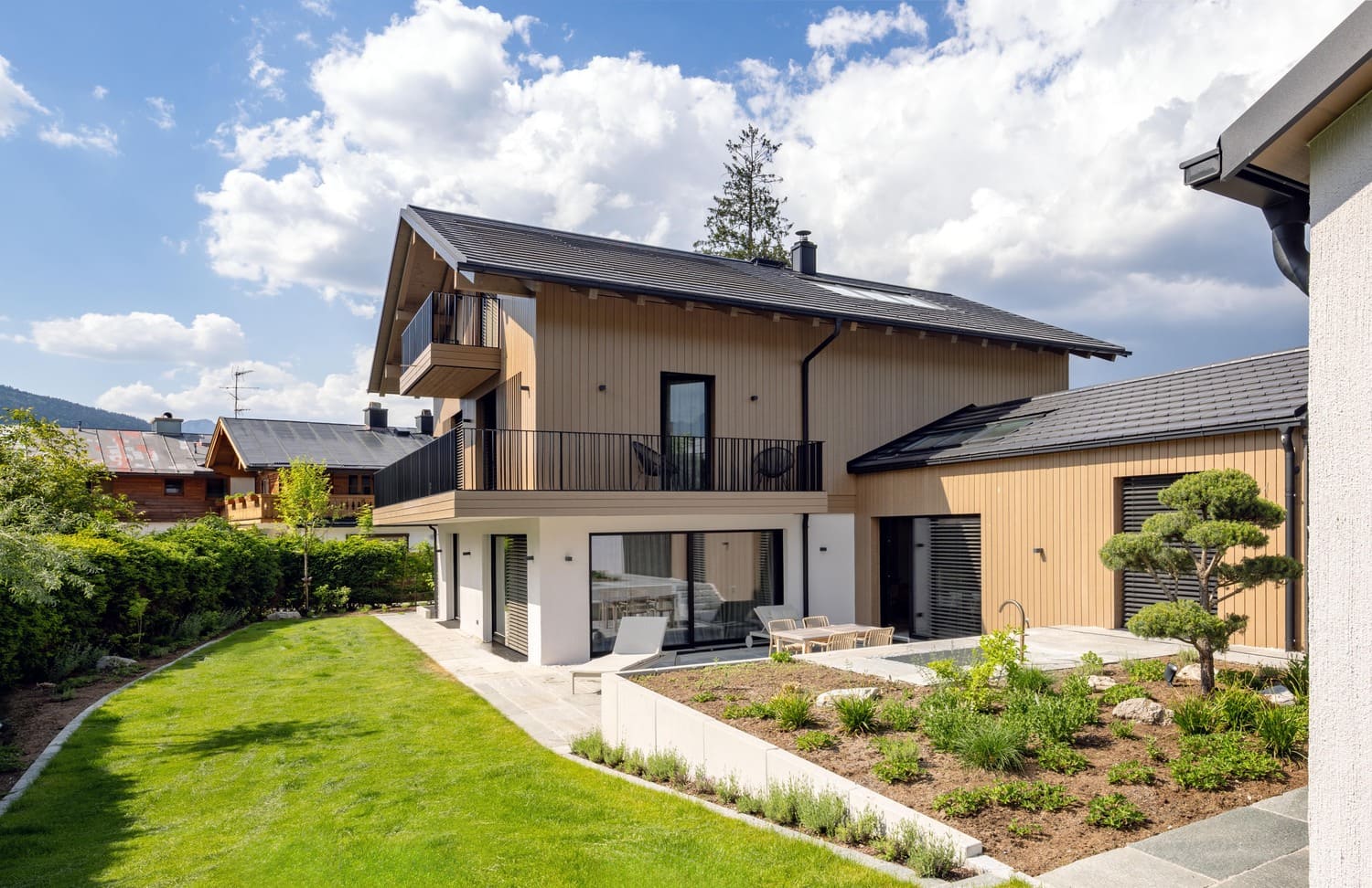 Modernes Haus mit Garten und Balkonen, umgeben von grüner Landschaft und blauem Himmel. | © Name