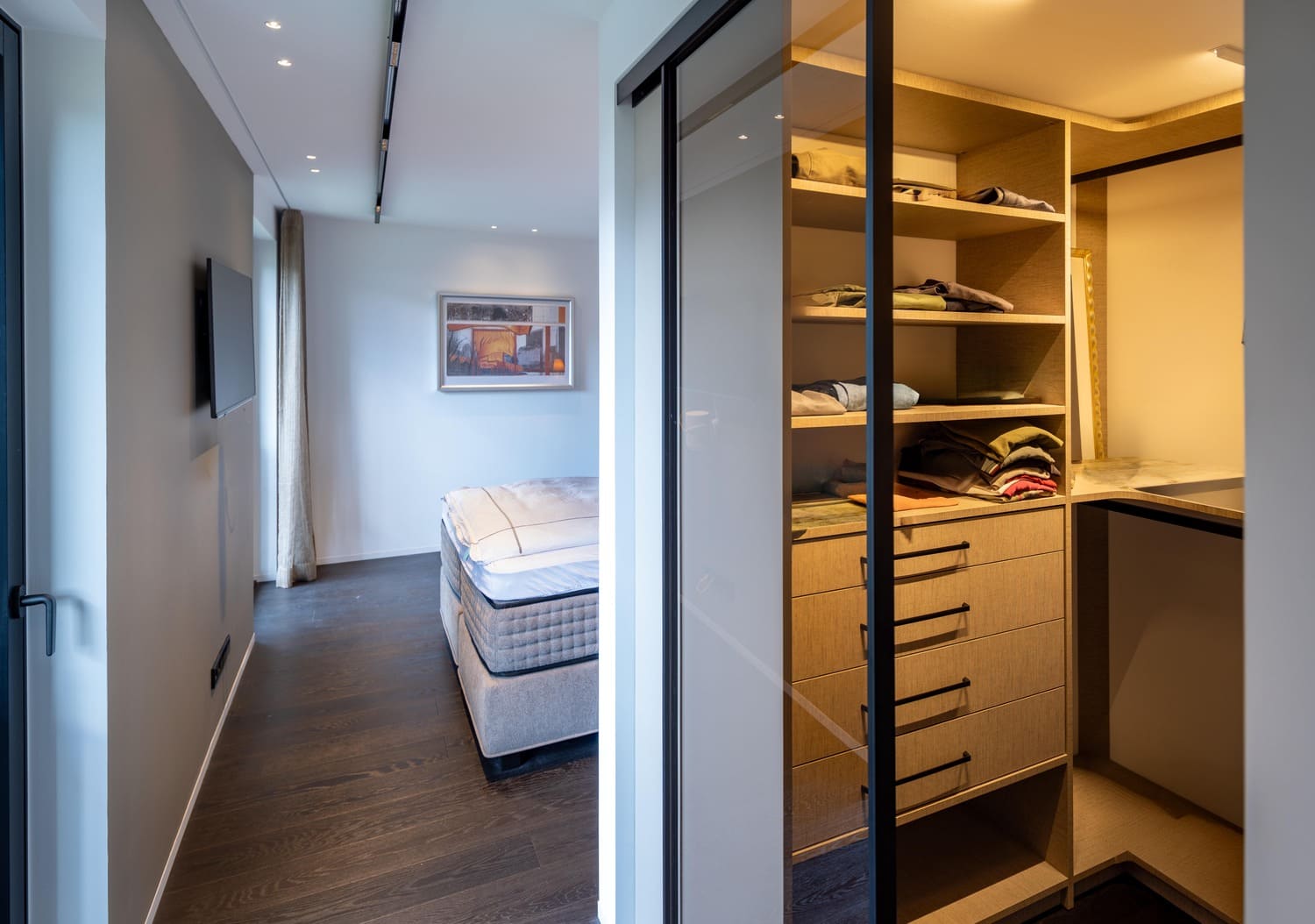 Ein moderner Kleiderschrank mit ordentlich gefalteter Kleidung und Blick auf ein Schlafzimmer mit Bett und TV.  | © Name