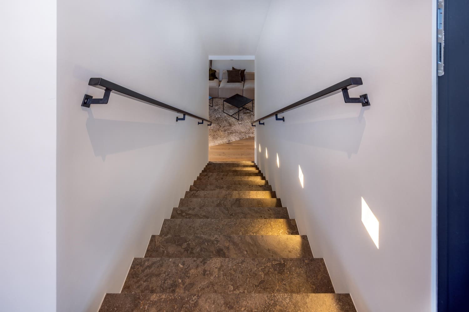 Eine helle Treppe mit Handlauf führt hinunter zu einem gemütlichen Wohnbereich im Hintergrund. | © Name