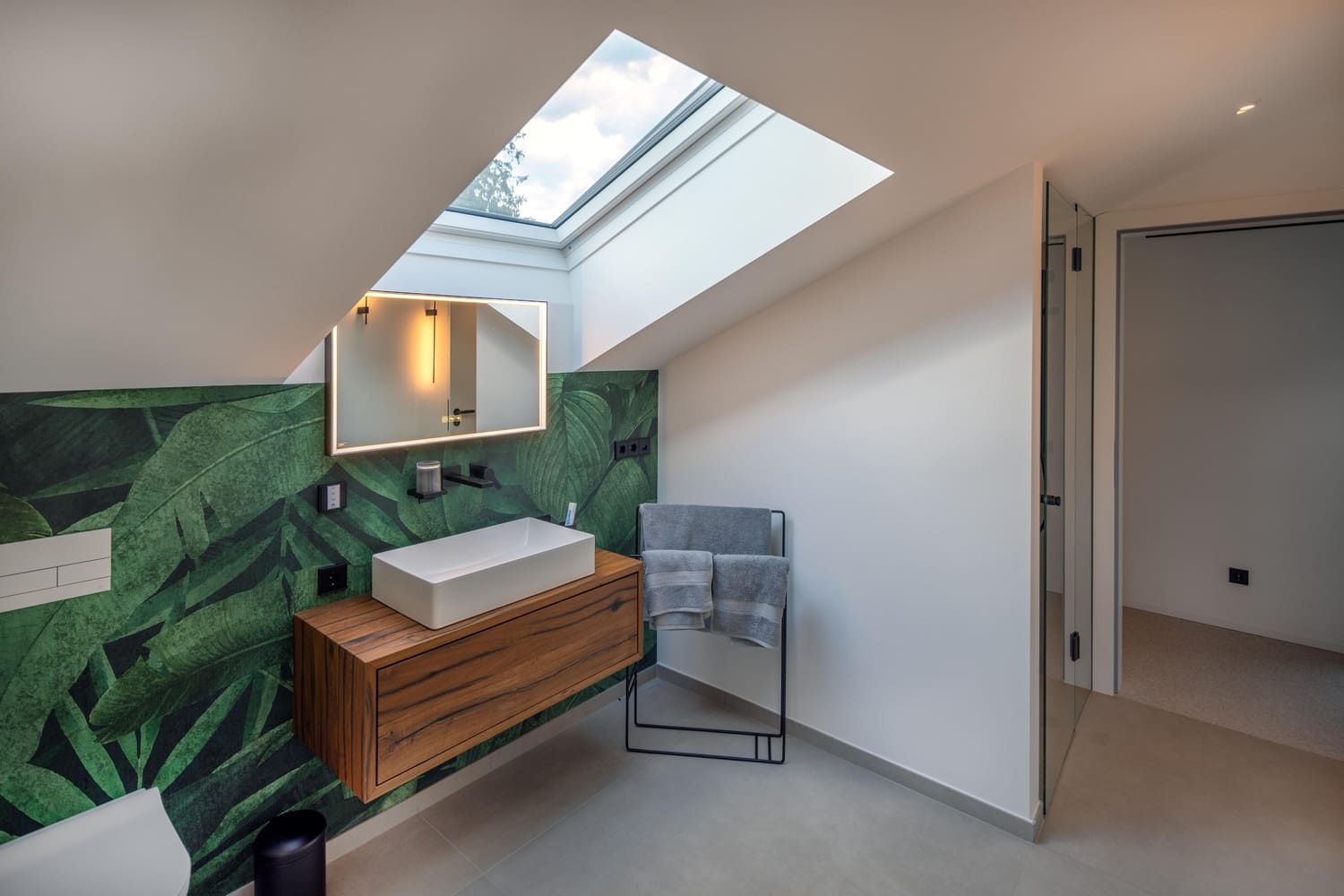 Modernes Badezimmer mit Holzwaschtisch, Pflanzenmuster und Dachfenster für natürliches Licht. | © Name