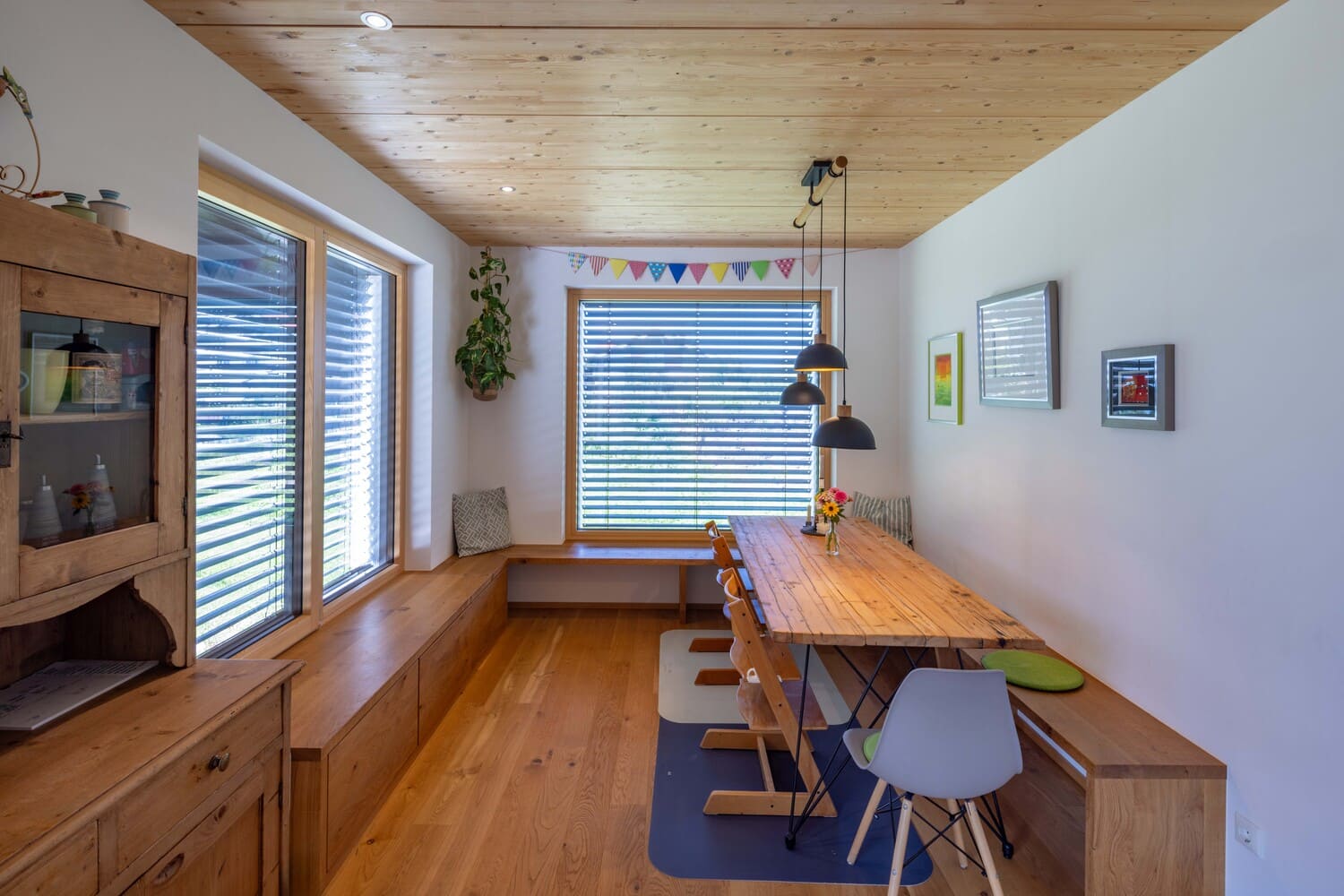 Ein gemütlicher Essbereich mit Holztisch und bunten Stühlen wird von Sonnenlicht durch große Fenster mit Jalousien erhellt.