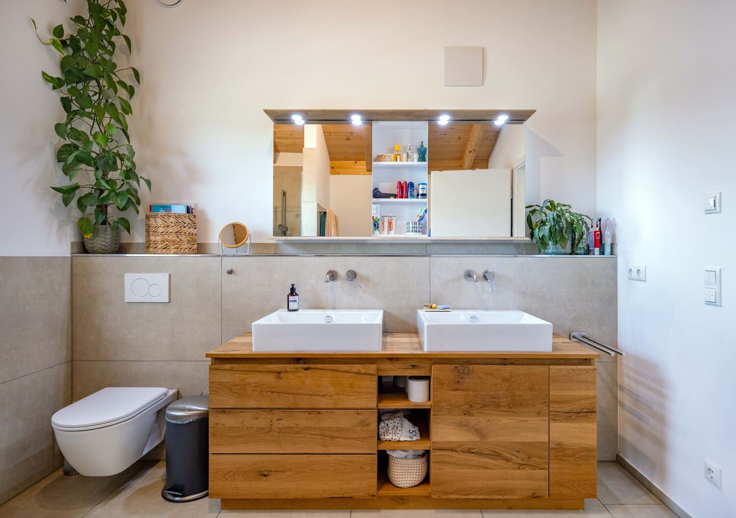 Modernes Badezimmer mit Doppelwaschbecken auf einem Holzmöbel, Pflanzen und Regalen unter einem großen, beleuchteten Spiegel.